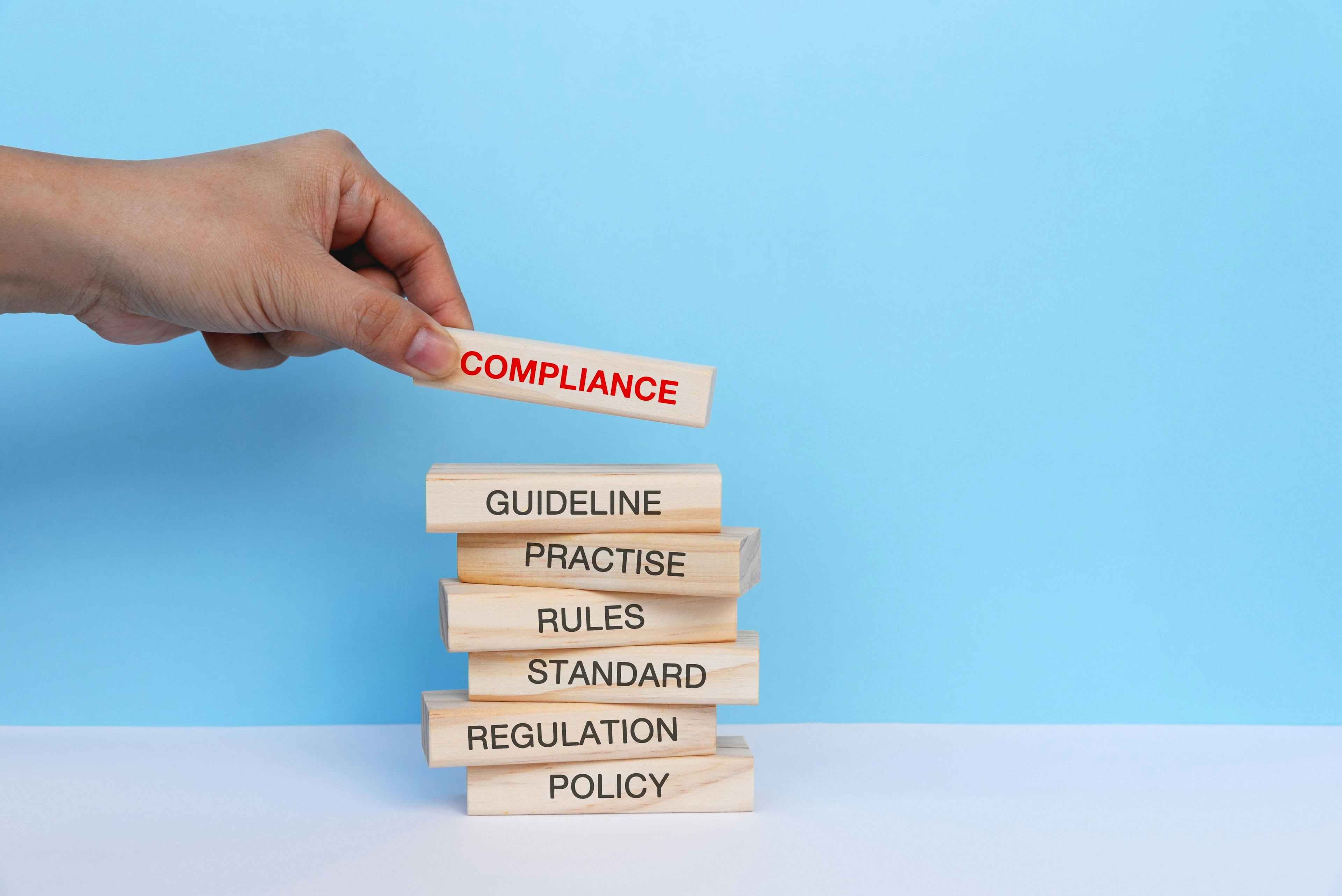 A hand holding out a long building block with "Compliance" text on it stacking it on a stack of blocks with "